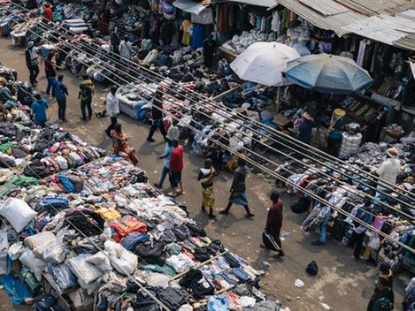 Markt mit mehreren Ständen, an denen Kleidung und andere Waren auf dem Boden und auf Tischen ausgebreitet sind, mit Menschen, die sich zwischen den Ständen bewegen.