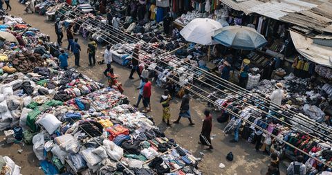 Market with multiple stalls displaying clothing and other goods spread on the ground and tables with people walking between the stalls.