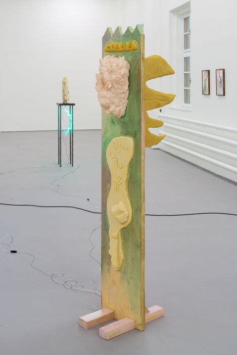 Sculpture made of painted wood with a relief face and yellow wings in a white gallery space.