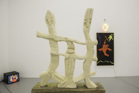 Sculpture of light-colored abstract figures with outstretched arms in front of a white wall with a small television and a glowing colorful artwork on the wall.