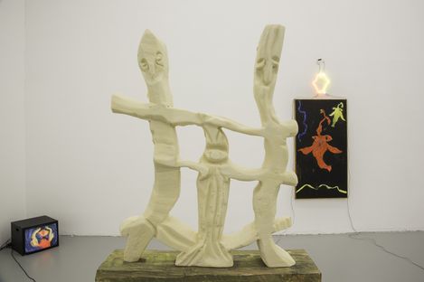 Sculpture of light-colored abstract figures with outstretched arms in front of a white wall with a small television and a glowing colorful artwork on the wall.