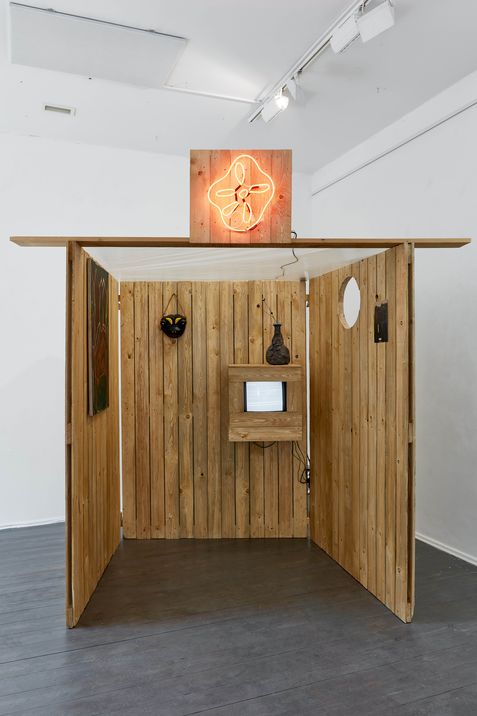 Wooden cabin with three walls and an open entrance, a small screen and a vase on the back wall, and a glowing star-shaped neon sign on the roof.