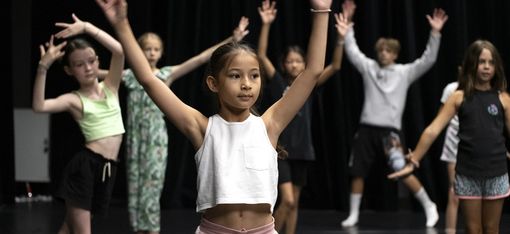 Kinder stehen mit einem zum V erhobenen Armen auf einem Tanzboden.