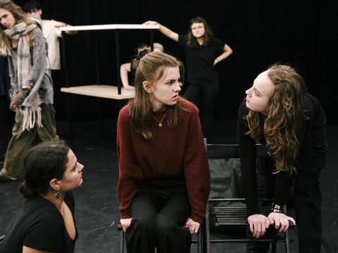 There are seven people on stage. In the foreground, two young women are looking at a third woman with reproachful, threatening expressions. In the background, four people are watching the scene.