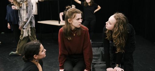 There are seven people on stage. In the foreground, two young women are looking at a third woman with reproachful, threatening expressions. In the background, four people are watching the scene.