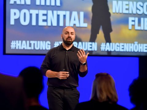 Ali Mahlodji is standing on a stage, speaking to the audience. Parts of a graphic overlay can be seen in the background.