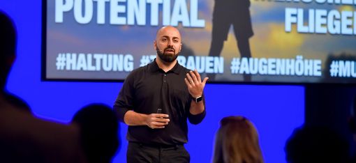 Ali Mahlodji is standing on a stage, speaking to the audience. Parts of a graphic overlay can be seen in the background.