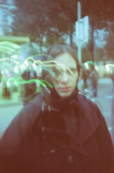 Portrait of a person wearing a dark coat with slicked-back hair against a blurred evening city background with lights.