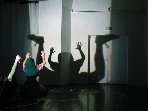 Two people are sitting on the floor, casting shadows onto a wall. Their shadows form the shape of a frog lying on its back.