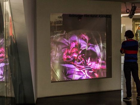 Person standing in front of a wall with a projection of abstract colorful shapes and the text "Animation Film Kunst ASIFAKEIL".