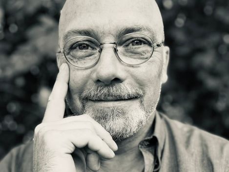 Porträt eines lächelnden, glatzköpfigen Mannes mit Brille, der mit der rechten Hand an die rechte Gesichtshälfte fasst.