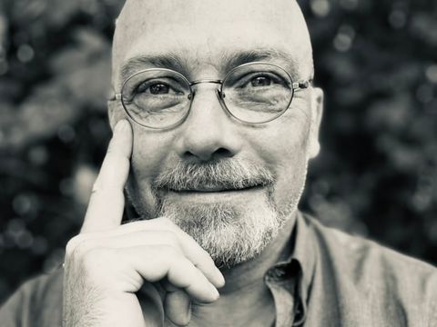 Portrait of a smiling bald man wearing glasses touching the right side of his face with his right hand.