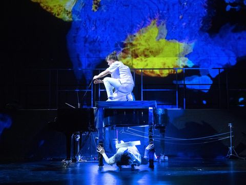 An impressive stage set is on display: a musician is sitting on a grand piano, with another musician beneath it. In the background, a pattern in blue and yellow is projected onto a huge screen.