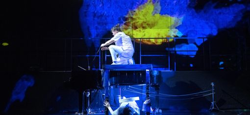 An impressive stage set is on display: a musician is sitting on a grand piano, with another musician beneath it. In the background, a pattern in blue and yellow is projected onto a huge screen.