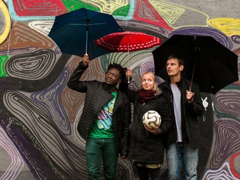 Three people are standing in front of a colourfully painted wall, holding umbrellas. The woman in the middle is holding a ball.