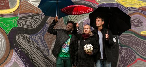 Three people are standing in front of a colourfully painted wall, holding umbrellas. The woman in the middle is holding a ball.