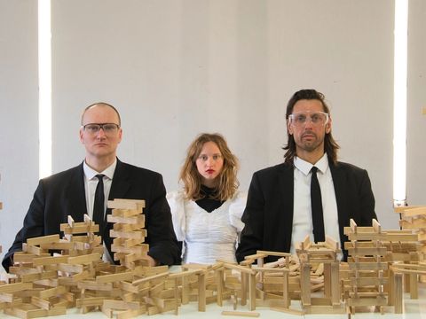 Drei Personen sehen mit strengem Blick und bestückt mit Laborbrillen in die Kamera. Vor ihnen stehen unzählige Holzbauklötze.