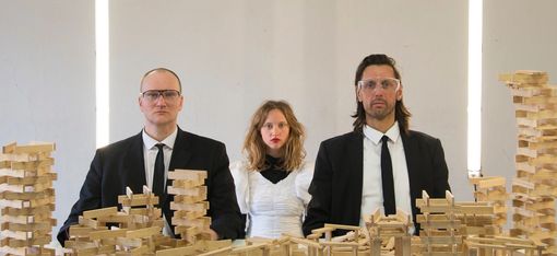 Drei Personen sehen mit strengem Blick und bestückt mit Laborbrillen in die Kamera. Vor ihnen stehen unzählige Holzbauklötze.