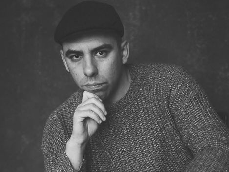 Black-and-white portrait of a seated person wearing a hat and sweater with the face obscured
