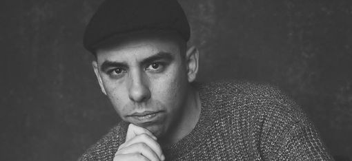 Black-and-white portrait of a seated person wearing a hat and sweater with the face obscured
