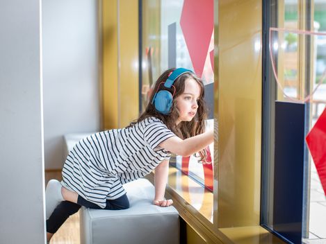 Ein Kind lehnt auf einem Sitzwürfel und hat Kopfhörer auf dem Kopf. Sie schaut aus dem Fenster.