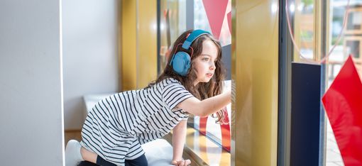 A child leans against a cube-shaped seat and wears headphones. She looks out the window.
