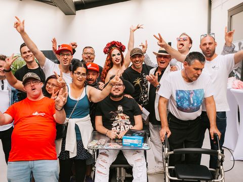 Gruppe von Menschen in einem Raum, einige machen Friedenszeichen, eine Person sitzt im Rollstuhl, eine weitere trägt ein rotes Kleid und Kopfschmuck