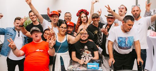 Gruppe von Menschen in einem Raum, einige machen Friedenszeichen, eine Person sitzt im Rollstuhl, eine weitere trägt ein rotes Kleid und Kopfschmuck