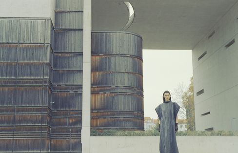 A person stands in front of a large rust-colored cylindrical metal building with a bright rectangular passage in the background