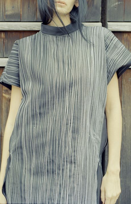 Upper body of a person wearing a black short-sleeve top with white vertical lines standing in front of a wooden wall