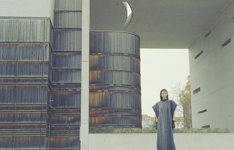A person stands in front of a large rust-colored cylindrical metal building with a bright rectangular passage in the background