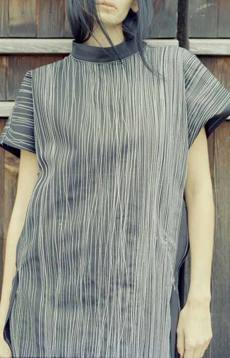 Upper body of a person wearing a black short-sleeve top with white vertical lines standing in front of a wooden wall