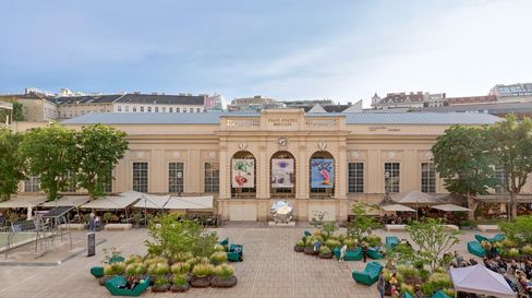 Außenhof des MuseumsQuartiers in Wien mit einem historischen Gebäude, an dem große Banner angebracht sind. Sitzbereiche mit Pflanzkübeln sorgen für eine entspannte Atmosphäre.