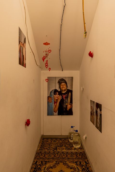 Narrow room with carpeted floor, photos of people on the walls, red heart decorations hanging from the ceiling, and two large water bottles on the floor.