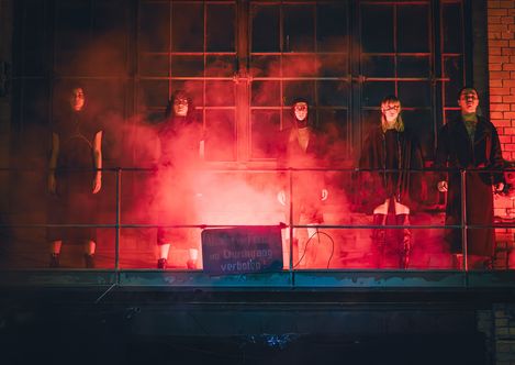 Five people stand side by side on a balcony in front of a large window surrounded by red smoke
