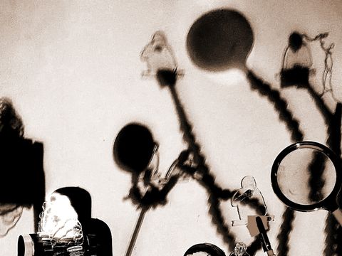 Verschwommene Schatten von Figuren und Objekten an einer Wand, darunter eine Lupe und mehrere Stäbe mit runden Enden