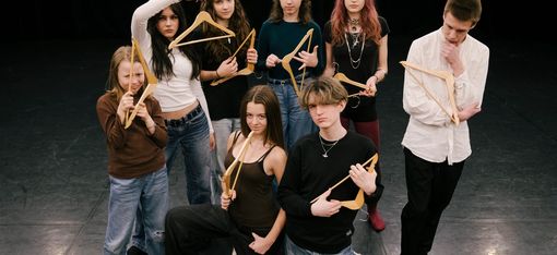Eight people are standing in an empty room. They are holding hangers in their hands. Their facial expressions are questioning and critical.