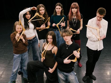 Eight people are standing in an empty room. They are holding hangers in their hands. Their facial expressions are questioning and critical.