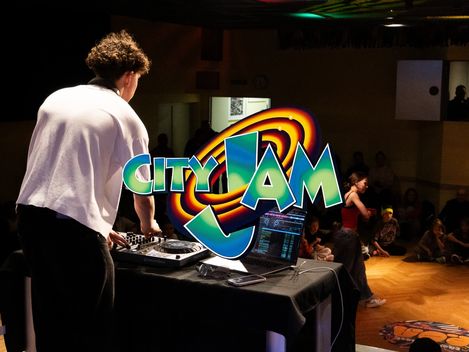 Eine Person ist von hinten an einem Mischpult stehend zu sehen. Kinder sitzen am Rande der Tanzfläche und beobachten eine Person, die auf die Fläche tritt. Der Schriftzug "City Jam" ist mittig auf dem Foto platziert.