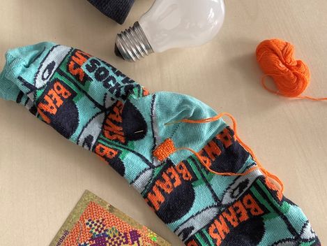 Zwei bunte Socken mit Schriftzug "BEAMS" liegen auf einem hellen Untergrund neben einer Glühbirne und einem kleinen Knäuel oranger Wolle.