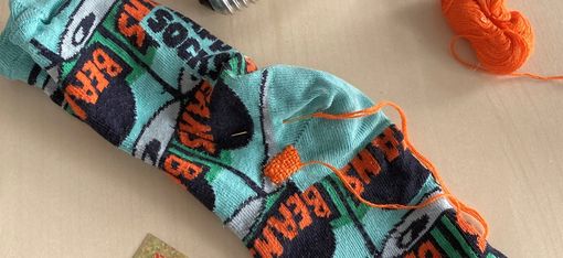 Two colorful socks with the word "BEAMS" lie on a light surface next to a light bulb and a small ball of orange yarn.
