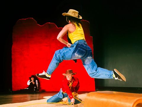 Two female cowboys in action: one is kneeling on the ground and swinging her lasso, whilst a second one leaps over her in a long stride. A third is leaning against a red wall in the background, watching the action unfold. All three are wearing impressive cowboy hats.
