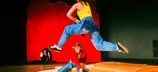 Two female cowboys in action: one is kneeling on the ground and swinging her lasso, whilst a second one leaps over her in a long stride. A third is leaning against a red wall in the background, watching the action unfold. All three are wearing impressive cowboy hats.