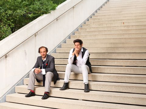 Zwei Männer sitzen auf einer breiten Treppe im Freien. Einer trägt ein dunkles Sakko mit Krawatte, der andere ein weißes Hemd mit schwarzer Weste.