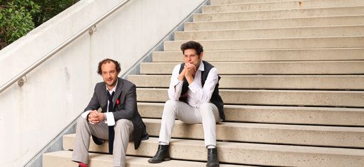 Zwei Männer sitzen auf einer breiten Treppe im Freien. Einer trägt ein dunkles Sakko mit Krawatte, der andere ein weißes Hemd mit schwarzer Weste.
