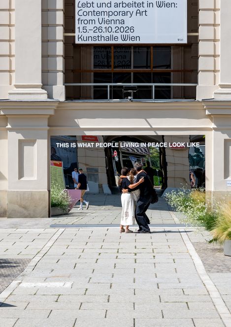 Zwei Personen umarmen sich vor einem großen Spiegel mit der Aufschrift 'THIS IS WHAT PEOPLE LIVING IN PEACE LOOK LIKE' vor einem historischen Gebäude mit einem Banner zur Ausstellung 'Lebt und arbeitet in Wien: Contemporary Art from Vienna 1.5.–26.10.2026 Kunsthalle Wien'