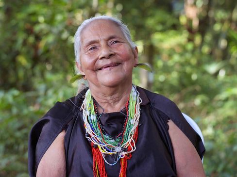 Person with white hair wearing multiple colorful bead necklaces and black clothing against a green background