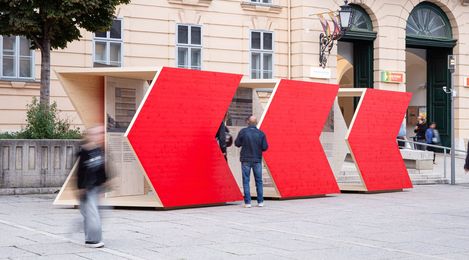 Three large red wooden arrow shapes arranged in a row on a plaza in front of a historic building