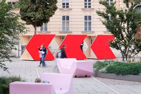 Three large red arrows on wooden panels in front of a multi-story building with trees and benches in the foreground