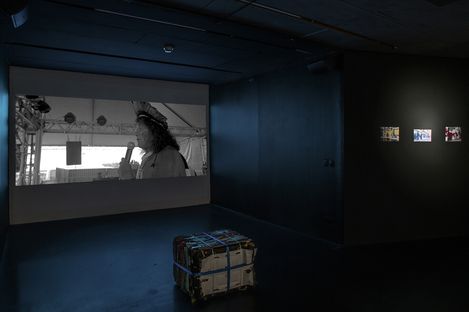 A dimly lit room features a large black and white projection of a speaker with a microphone and headdress. Three small, illuminated artworks are displayed on the right wall, and a bundled, taped cube sits on the dark floor, setting a contemplative tone.
